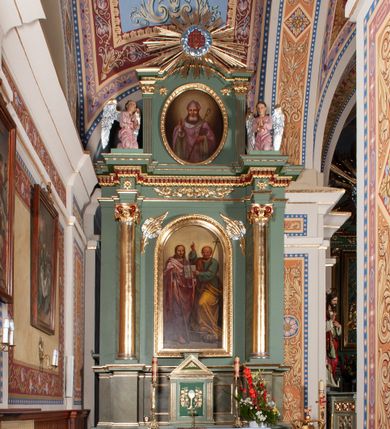 Zdjęcie nr 1: Ołtarz architektoniczny, jednoosiowy, jednokondygnacyjny ze zwieńczeniem. Mensa ołtarzowa prostopadłościenna, antepedium ujęte w ramę z dekoracją ornamentalną z greckim krzyżem pośrodku. Tabernakulum w formie prostopadłościennej skrzynki, ujęte po bokach parą kanelowanych pilastrów, wspierających profilowany gzyms i trójkątny przyczółek, za którym stoi prosty tron wystawienia w formie podwyższonego cokołu. Drzwiczki w kształcie stojącego prostokąta z wpisaną w polu złoconą dekoracją winnej latorośli i motywem kielicha z hostią. Retabulum ołtarzowe wsparte na dwustrefowym cokole, ujęte po bokach parą zdwojonych pilastrów. Kondygnacja flankowana parą złoconych kolumn z kompozytowymi kapitelami, ustawionych na tle pilastrów. Podpory wspierają pełne belkowanie wyłamane nad polem środkowym. Na osi kolumn usadzona jest para klęczących aniołów. W polu głównym obraz w kształcie stojącego prostokąta zakończonego łukiem półkolistym przedstawiający świętych apostołów: Filipa i Jakuba. W górnych przyłuczach pola środkowego para gałązek palmowych przeplecionych przez wianki. Fryz zdobiony wicią z liści akantu, na impostach rozetki, wyżej czerwony gzyms dekorowany kostkowaniem i kimationem. Zwieńczenie w kształcie stojącego prostokąta, flankowane parą kanelowanych pilastrów o korynckich kapitelach podtrzymujących wyłamany gzyms w kształcie łuku nadwieszonego. W polu owalny obraz św. Mikołaja. U szczytu zatknięta promienista gloria z wpisanym pośrodku monogramem IHS na czerwonym tle w wieńcu utworzonym z obłoków. Struktura polichromowana w kolorze jasnozielonym, detale, ornamentyka i profilowania złocone i srebrzone.