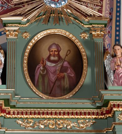 Zdjęcie nr 2: Obraz w kształcie owalu z przedstawieniem św. Mikołaja. Święty ukazany w półpostaci, frontalnie, z prawą ręką wzniesioną w geście błogosławieństwa, z pastorałem w lewej, w której przytrzymuje również księgę. Twarz szeroka o wyraźnie podkreślonych kościach policzkowych, oczy wzniesione ku górze, broda siwa i gęsta. Ubrany jest w białą albę, różowy i szeroki ornat z zieloną podszewką oraz biały paliusz. Na głowie ma założoną fioletową infułę, zdobioną prostym krzyżykiem. Pastorał o prostych formach z zawiniętą krzywaśnią, bez zdobień. Księga w czerwonej oprawie z napisem „S(więty) Jan / Chryzostom / [...]”. Tło jednolite, ciemnobrązowe, rozjaśnione wokół głowy świętego. Rama drewniana, złocona, dekorowana kimationem.

