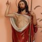 Zdjęcie nr 1: Rzeźba Chrystusa Zmartwychwstałego pełnoplastyczna, wolnostojąca. Chrystus ukazany w całej postaci ustawiony frontalnie, w delikatnym kontrapoście, prawą ręką uniesioną na wysokość głowy czyni gest błogosławieństwa, w lewej trzyma chorągiew o długim drzewcu, z białym krzyżem na czerwonym płacie. Chrystus ma pociągłą twarz, z długim i wąskim nosem, wzrokiem skierowanym w dal, okoloną krótką i ciemną brodą i długimi oraz falowanymi włosami, które opadają na plecy i ramiona. Wokół głowy ma okrągły nimb w formie złoconej obręczy z drutu. Jest ubrany w czerwony płaszcz z białą podszewką, dekorowany ornamentem roślinnym, spięty pod szyją broszą, odsłaniający nagi tors, otaczający figurę i przerzucony jedną połą przez lewe ramię. Na dłoniach, bosych stopach i boku zaznaczone są ślady męki. Polichromia ciała jest naturalistyczna.