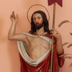 Zdjęcie nr 1: Rzeźba Chrystusa Zmartwychwstałego pełnoplastyczna, wolnostojąca. Chrystus ukazany w całej postaci ustawiony frontalnie, w delikatnym kontrapoście, prawą ręką uniesioną na wysokość głowy czyni gest błogosławieństwa, w lewej trzyma chorągiew o długim drzewcu, z białym krzyżem na czerwonym płacie. Chrystus ma pociągłą twarz, z długim i wąskim nosem, wzrokiem skierowanym w dal, okoloną krótką i ciemną brodą i długimi oraz falowanymi włosami, które opadają na plecy i ramiona. Wokół głowy ma okrągły nimb w formie złoconej obręczy z drutu. Jest ubrany w czerwony płaszcz z białą podszewką, dekorowany ornamentem roślinnym, spięty pod szyją broszą, odsłaniający nagi tors, otaczający figurę i przerzucony jedną połą przez lewe ramię. Na dłoniach, bosych stopach i boku zaznaczone są ślady męki. Polichromia ciała jest naturalistyczna.