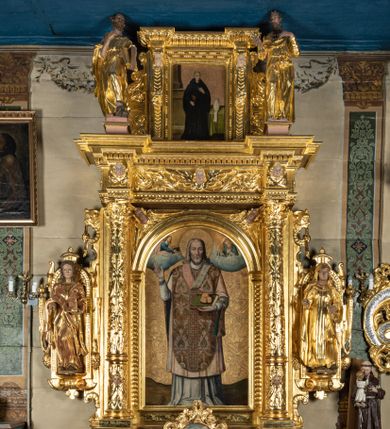 Zdjęcie nr 1: Ołtarz przyścienny, dwukondygnacyjny, jednoosiowy o formach architektonicznych; uszaty. Mensa drewniana, prostopadłościenna; antependium ujęte płaskorzeźbioną, złoconą ramą z dekoracją akantową. Lico malowane z monogramem maryjnym w otoczeniu barwnej dekoracji roślinno-kwiatowej na jasnobeżowym tle. W predelli rama w kształcie leżącego prostokąta ujęta ornamentem kartuszowym mieszcząca pierwotnie niezachowaną dekorację malarską. Nastawa ustawiona na wysokim cokole, ujęta kompozytowymi kolumnami dźwigającymi przełamujące się na osiach podpór pełne belkowanie z kostkowanym achitrawem, płaskorzeźbionym w motyw akantu i uskrzydlonych główek anielskich fryzem oraz wydatnym gzymsem. Kolumny w jednej trzeciej wysokości ujęte profilowanymi opaskami; trzony poniżej opasek zdobione motywem główek anielskich; powyżej płaskorzeźbiona dekoracja arabeskowa. W polu ołtarza prostokątna rama zamknięta łukiem półokrągłym mieszcząca obraz św. Mikołaja; w przyłuczach ramy umieszczone płaskorzeźbione, uskrzydlone główki anielskie. Kondygnacja druga ujęta hermowymi pilastrami dźwigającymi uproszczone belkowanie z wydatnym gzymsem. W polu obraz św. Benedykta o kształcie stojącego prostokąta w złoconej, profilowanej ramie. Na osiach kolumn, ustawione rzeźby św. Piotra (z prawej) i św. Pawła (z lewej) na tle esowatych wolut. Całość nastawy ujęta ażurowymi uszami utworzonymi z ornamentu okuciowego mieszczącymi nisze z rzeźbami św. Cecylii (z lewej) oraz św. Teresy z Avila (z prawej) umieszczonymi na tle muszli. Ponad niszami maszkarony trzymające w pyskach spływające do dołu draperie. Struktura ołtarza oraz partie szat postaci złocone; karnacje rzeźb polichromowane.
