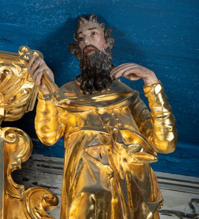 Zdjęcie nr 18: Ołtarz przyścienny, dwukondygnacyjny, jednoosiowy o formach architektonicznych; uszaty. Mensa drewniana, prostopadłościenna; antependium ujęte płaskorzeźbioną, złoconą ramą z dekoracją akantową. Lico malowane z monogramem maryjnym w otoczeniu barwnej dekoracji roślinno-kwiatowej na jasnobeżowym tle. W predelli rama w kształcie leżącego prostokąta ujęta ornamentem kartuszowym mieszcząca pierwotnie niezachowaną dekorację malarską. Nastawa ustawiona na wysokim cokole, ujęta kompozytowymi kolumnami dźwigającymi przełamujące się na osiach podpór pełne belkowanie z kostkowanym achitrawem, płaskorzeźbionym w motyw akantu i uskrzydlonych główek anielskich fryzem oraz wydatnym gzymsem. Kolumny w jednej trzeciej wysokości ujęte profilowanymi opaskami; trzony poniżej opasek zdobione motywem główek anielskich; powyżej płaskorzeźbiona dekoracja arabeskowa. W polu ołtarza prostokątna rama zamknięta łukiem półokrągłym mieszcząca obraz św. Mikołaja; w przyłuczach ramy umieszczone płaskorzeźbione, uskrzydlone główki anielskie. Kondygnacja druga ujęta hermowymi pilastrami dźwigającymi uproszczone belkowanie z wydatnym gzymsem. W polu obraz św. Benedykta o kształcie stojącego prostokąta w złoconej, profilowanej ramie. Na osiach kolumn, ustawione rzeźby św. Piotra (z prawej) i św. Pawła (z lewej) na tle esowatych wolut. Całość nastawy ujęta ażurowymi uszami utworzonymi z ornamentu okuciowego mieszczącymi nisze z rzeźbami św. Cecylii (z lewej) oraz św. Teresy z Avila (z prawej) umieszczonymi na tle muszli. Ponad niszami maszkarony trzymające w pyskach spływające do dołu draperie. Struktura ołtarza oraz partie szat postaci złocone; karnacje rzeźb polichromowane.
