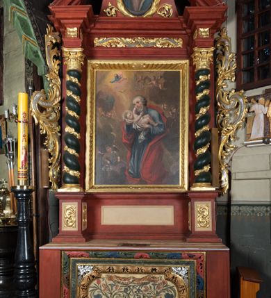 Zdjęcie nr 1: Ołtarz przyścienny o formach architektonicznych, jednoosiowy, jednokondygnacyjny ze zwieńczeniem; ujęty akantowymi uszami. Mensa prostopadłościenna; antepedium malowane w symetryczne motywy roślinno-kwiatowe na jasnobeżowym tle; ujęte złoconą ramą w kształcie leżącego prostokąta o wypukłych krótszych bokach. Nastawa ujęta parą kolumn umieszczonych na wysokich, prostopadłościennych postumentach. Trzony kolumn skręcone, oplecione kwiatowymi girlandami; kapitele kompozytowe, złocone. W polu ołtarza złocona, profilowana rama w kształcie stojącego prostokąta, mieszcząca obraz z przedstawieniem św. Józefa. Kolumny dźwigają pełne belkowanie przełamane na osiach podpór oraz odcinki trójkątnego przyczółka, na których pełnoplastyczne figurki aniołów. W partii fryzu złocony ornament akantowy oraz rozetki. Zwieńczenie w kształcie równoramiennego trapezu o wklęsłych bokach, zamknięte od góry gzymsem wklęsło-wypukłym. W polu owalna, złocona rama w formie wieńca laurowego mieszcząca obraz przedstawiający św. Jana Ewangelistę, ponad którym owalny, akantowy kartusz z napisem „IN / HONOREM S(ANCTI) JOSEPHI / ANNO DOMINI 1528”. Nastawa ujęta ażurowymi, złoconymi i srebrzonymi uszami utworzonymi z suchego akantu. Struktura ołtarza marmoryzowana w odcieniach czerwieni; dekoracja snycerska srebrzona i złocona, trzony kolumn czarne.
