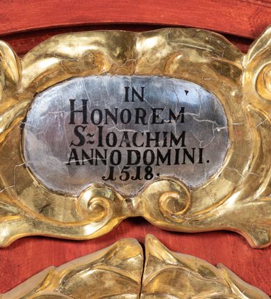 Zdjęcie nr 3: Ołtarz przyścienny o formach architektonicznych; jednoosiowy, jednokondygnacyjny ze zwieńczeniem, uszaty. Mensa prostopadłościenna; antepedium malowane z trzema kwiatowymi medalionami mieszczącymi literę J, monogram maryjny oraz literę A na jasnobeżowym tle. Poniżej środkowego medalionu płonące serce przebite krzyżem i kotwicą. W predelli nisza w kształcie poziomego prostokąta z wypukłymi krótszymi bokami, zapewne pierwotnie mieszcząca niezachowany obraz. Nastawa ujęta parą kolumn umieszczonych na wysokich, prostopadłościennych postumentach. Trzony kolumn skręcone, oplecione kwiatowymi girlandami; kompozytowe kapitele złocone. W polu ołtarza złocona, profilowana rama w kształcie pionowego prostokąta mieszcząca obraz z przedstawieniem św. Anny, św. Joachima i Marii. Kolumny dźwigają pełne belkowanie przełamane na osiach podpór oraz odcinki zbudowanego z wolut przyczółka, na których umieszczono pełnoplastyczne figurki aniołów. W partii fryzu złocony ornament akantowy przepleciony srebrzoną wstęgą  oraz rozetki. Zwieńczenie w kształcie trapezu równoramiennego o wklęsłych bokach, zamknięte od góry gzymsem w formie łuku nadwieszonego. W polu owalna, złocona rama w kształcie wieńca laurowego mieszcząca obraz przedstawiający św. Sebastiana, ponad którym akantowy kartusz owalny z inskrypcją „IN HONOREM S(ANCTI) JOACHIM ANNO DOMINI 1518”. Nastawa ujęta ażurowymi, złoconymi i srebrzonymi uszami utworzonymi z suchego akantu oraz karbowanej wstęgi. Struktura ołtarza „marmoryzowana” w odcieniach czerwieni; dekoracja snycerska srebrzona i złocona, trzony kolumn czarne.



