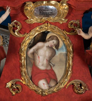 Zdjęcie nr 14: Ołtarz przyścienny o formach architektonicznych; jednoosiowy, jednokondygnacyjny ze zwieńczeniem, uszaty. Mensa prostopadłościenna; antepedium malowane z trzema kwiatowymi medalionami mieszczącymi literę J, monogram maryjny oraz literę A na jasnobeżowym tle. Poniżej środkowego medalionu płonące serce przebite krzyżem i kotwicą. W predelli nisza w kształcie poziomego prostokąta z wypukłymi krótszymi bokami, zapewne pierwotnie mieszcząca niezachowany obraz. Nastawa ujęta parą kolumn umieszczonych na wysokich, prostopadłościennych postumentach. Trzony kolumn skręcone, oplecione kwiatowymi girlandami; kompozytowe kapitele złocone. W polu ołtarza złocona, profilowana rama w kształcie pionowego prostokąta mieszcząca obraz z przedstawieniem św. Anny, św. Joachima i Marii. Kolumny dźwigają pełne belkowanie przełamane na osiach podpór oraz odcinki zbudowanego z wolut przyczółka, na których umieszczono pełnoplastyczne figurki aniołów. W partii fryzu złocony ornament akantowy przepleciony srebrzoną wstęgą  oraz rozetki. Zwieńczenie w kształcie trapezu równoramiennego o wklęsłych bokach, zamknięte od góry gzymsem w formie łuku nadwieszonego. W polu owalna, złocona rama w kształcie wieńca laurowego mieszcząca obraz przedstawiający św. Sebastiana, ponad którym akantowy kartusz owalny z inskrypcją „IN HONOREM S(ANCTI) JOACHIM ANNO DOMINI 1518”. Nastawa ujęta ażurowymi, złoconymi i srebrzonymi uszami utworzonymi z suchego akantu oraz karbowanej wstęgi. Struktura ołtarza „marmoryzowana” w odcieniach czerwieni; dekoracja snycerska srebrzona i złocona, trzony kolumn czarne.



