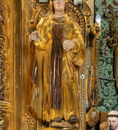 Zdjęcie nr 1: Rzeźba drążona, ustawiona na niewielkim prostopadłościennej podstawie na tle niszy w prawym naddatku ołtarza. Przedstawia św. Teresę z Avila ukazaną frontalnie, w pozycji stojącej, w lekkim kontrapoście z lewą nogą ugiętą w kolanie i wysunięta do przodu. Święta została przedstawiona jako dojrzała kobieta o owalnej twarzy z pełnymi policzkami, wyraźnie zaznaczonym niewielkim podbródkiem, wydatnym prostym nosem oraz migdałowatymi, wpatrzonymi na wprost oczami. Włosy niewidoczne, zakryte welonem zakonnym. Prawa ręka postaci ugięta w łokciu i wysunięta do przodu. Układ palców prawej dłoni wskazuje, że być może w tym miejscu postać trzymała atrybut, który nie zachował się do dziś. Lewa ręka również ugięta w łokciu i swobodnie wysunięta przed siebie. Święta jest ubrana w obszerny złoty habit przepasany na wysokości talii, na którym widoczny szkaplerz oraz płaszcz. Z lewej strony poła płaszcza swobodnie opada w dół, z prawej tkanina tworzy łyżkowatą fałdę umocowaną przy pasku habitu. Spod habitu widoczne czubki brązowych butów o zaokrąglonych noskach. Partie karnacji polichromowane, szaty w całości złocone.
