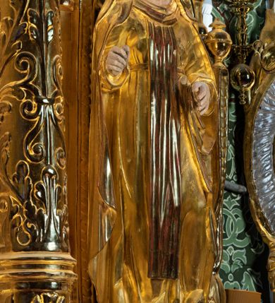 Zdjęcie nr 3: Rzeźba drążona, ustawiona na niewielkim prostopadłościennej podstawie na tle niszy w prawym naddatku ołtarza. Przedstawia św. Teresę z Avila ukazaną frontalnie, w pozycji stojącej, w lekkim kontrapoście z lewą nogą ugiętą w kolanie i wysunięta do przodu. Święta została przedstawiona jako dojrzała kobieta o owalnej twarzy z pełnymi policzkami, wyraźnie zaznaczonym niewielkim podbródkiem, wydatnym prostym nosem oraz migdałowatymi, wpatrzonymi na wprost oczami. Włosy niewidoczne, zakryte welonem zakonnym. Prawa ręka postaci ugięta w łokciu i wysunięta do przodu. Układ palców prawej dłoni wskazuje, że być może w tym miejscu postać trzymała atrybut, który nie zachował się do dziś. Lewa ręka również ugięta w łokciu i swobodnie wysunięta przed siebie. Święta jest ubrana w obszerny złoty habit przepasany na wysokości talii, na którym widoczny szkaplerz oraz płaszcz. Z lewej strony poła płaszcza swobodnie opada w dół, z prawej tkanina tworzy łyżkowatą fałdę umocowaną przy pasku habitu. Spod habitu widoczne czubki brązowych butów o zaokrąglonych noskach. Partie karnacji polichromowane, szaty w całości złocone.
