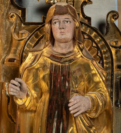 Zdjęcie nr 5: Rzeźba drążona, ustawiona na niewielkim prostopadłościennej podstawie na tle niszy w prawym naddatku ołtarza. Przedstawia św. Teresę z Avila ukazaną frontalnie, w pozycji stojącej, w lekkim kontrapoście z lewą nogą ugiętą w kolanie i wysunięta do przodu. Święta została przedstawiona jako dojrzała kobieta o owalnej twarzy z pełnymi policzkami, wyraźnie zaznaczonym niewielkim podbródkiem, wydatnym prostym nosem oraz migdałowatymi, wpatrzonymi na wprost oczami. Włosy niewidoczne, zakryte welonem zakonnym. Prawa ręka postaci ugięta w łokciu i wysunięta do przodu. Układ palców prawej dłoni wskazuje, że być może w tym miejscu postać trzymała atrybut, który nie zachował się do dziś. Lewa ręka również ugięta w łokciu i swobodnie wysunięta przed siebie. Święta jest ubrana w obszerny złoty habit przepasany na wysokości talii, na którym widoczny szkaplerz oraz płaszcz. Z lewej strony poła płaszcza swobodnie opada w dół, z prawej tkanina tworzy łyżkowatą fałdę umocowaną przy pasku habitu. Spod habitu widoczne czubki brązowych butów o zaokrąglonych noskach. Partie karnacji polichromowane, szaty w całości złocone.
