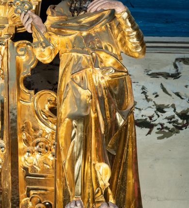 Zdjęcie nr 3: Rzeźba pełnoplastyczna ustawiona na niewielkim prostopadłościennym postumencie zwieńczonym złoconym gzymsem. Święty Piotr ukazany frontalnie w pozycji stojącej, w lekkim kontrapoście z prawą noga lekko ugiętą w kolanie i wysuniętą do przodu. Głowa zwrócona w trzech czwartych w lewo w kierunku osi ołtarza. Postać przedstawiona jako dojrzały mężczyzna o szczupłej, pociągłej twarzy okolonej ciemnym zarostem – wąsami i długą kędzierzawą brodą; włosy krótkie, ciemne i kręcone. Ręce zgięte w łokciach, przedramiona podniesione do góry. W prawej dłoni święty trzyma dwa klucze, lewa dłoń luźno opadająca, skierowana w lewo. Święty jest ubrany w długą, obficie fałdowaną, przewiązaną w pasie złotą szatę z długimi rękawami. Spod szaty wystają bose stopy. Partie karnacji polichromowane, szaty i atrybut złocone.