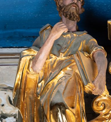 Zdjęcie nr 1: Rzeźba pełnoplastyczna ustawiona na niewielkim prostopadłościennym postumencie zwieńczonym złoconym gzymsem. Święty Paweł (?) ukazany frontalnie w pozycji stojącej, w wyraźnym kontrapoście z lewą noga lekko ugiętą w kolanie, wysuniętą do przodu i wspartą na skale. Głowa zwrócona w trzech czwartych w prawo w kierunku osi ołtarza. Włosy krótkie, kędzierzawe, przerzedzone nad czołem; zarost krótki, falowany. Twarz pociągła o wypukłym czole, oczy głęboko osadzone, nos prosty. Prawa ręka zgięta w łokciu i uniesiona do góry. Dłoń zwinięta do środka w geście trzymania – zapewne w tym miejscu znajdował się niezachowany atrybut. Lewa ręka opuszczona w dół, podtrzymująca dużą księgę. Święty ubrany w długą, obficie fałdowaną, przewiązaną w pasie złotą szatę z sięgającymi łokci rękawami oraz obszerny, bogato fałdowany płaszcz. Spod szaty wystają bose stopy. Partie karnacji polichromowane, szaty i atrybut złocone.


