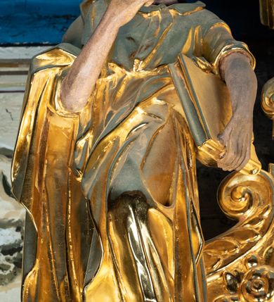 Zdjęcie nr 2: Rzeźba pełnoplastyczna ustawiona na niewielkim prostopadłościennym postumencie zwieńczonym złoconym gzymsem. Święty Paweł (?) ukazany frontalnie w pozycji stojącej, w wyraźnym kontrapoście z lewą noga lekko ugiętą w kolanie, wysuniętą do przodu i wspartą na skale. Głowa zwrócona w trzech czwartych w prawo w kierunku osi ołtarza. Włosy krótkie, kędzierzawe, przerzedzone nad czołem; zarost krótki, falowany. Twarz pociągła o wypukłym czole, oczy głęboko osadzone, nos prosty. Prawa ręka zgięta w łokciu i uniesiona do góry. Dłoń zwinięta do środka w geście trzymania – zapewne w tym miejscu znajdował się niezachowany atrybut. Lewa ręka opuszczona w dół, podtrzymująca dużą księgę. Święty ubrany w długą, obficie fałdowaną, przewiązaną w pasie złotą szatę z sięgającymi łokci rękawami oraz obszerny, bogato fałdowany płaszcz. Spod szaty wystają bose stopy. Partie karnacji polichromowane, szaty i atrybut złocone.


