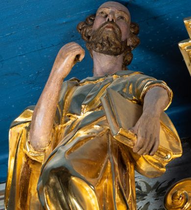 Zdjęcie nr 3: Rzeźba pełnoplastyczna ustawiona na niewielkim prostopadłościennym postumencie zwieńczonym złoconym gzymsem. Święty Paweł (?) ukazany frontalnie w pozycji stojącej, w wyraźnym kontrapoście z lewą noga lekko ugiętą w kolanie, wysuniętą do przodu i wspartą na skale. Głowa zwrócona w trzech czwartych w prawo w kierunku osi ołtarza. Włosy krótkie, kędzierzawe, przerzedzone nad czołem; zarost krótki, falowany. Twarz pociągła o wypukłym czole, oczy głęboko osadzone, nos prosty. Prawa ręka zgięta w łokciu i uniesiona do góry. Dłoń zwinięta do środka w geście trzymania – zapewne w tym miejscu znajdował się niezachowany atrybut. Lewa ręka opuszczona w dół, podtrzymująca dużą księgę. Święty ubrany w długą, obficie fałdowaną, przewiązaną w pasie złotą szatę z sięgającymi łokci rękawami oraz obszerny, bogato fałdowany płaszcz. Spod szaty wystają bose stopy. Partie karnacji polichromowane, szaty i atrybut złocone.


