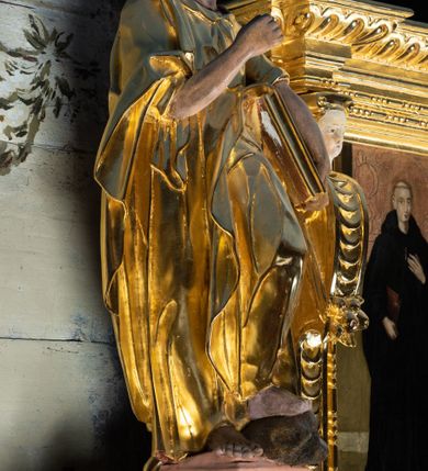 Zdjęcie nr 5: Rzeźba pełnoplastyczna ustawiona na niewielkim prostopadłościennym postumencie zwieńczonym złoconym gzymsem. Święty Paweł (?) ukazany frontalnie w pozycji stojącej, w wyraźnym kontrapoście z lewą noga lekko ugiętą w kolanie, wysuniętą do przodu i wspartą na skale. Głowa zwrócona w trzech czwartych w prawo w kierunku osi ołtarza. Włosy krótkie, kędzierzawe, przerzedzone nad czołem; zarost krótki, falowany. Twarz pociągła o wypukłym czole, oczy głęboko osadzone, nos prosty. Prawa ręka zgięta w łokciu i uniesiona do góry. Dłoń zwinięta do środka w geście trzymania – zapewne w tym miejscu znajdował się niezachowany atrybut. Lewa ręka opuszczona w dół, podtrzymująca dużą księgę. Święty ubrany w długą, obficie fałdowaną, przewiązaną w pasie złotą szatę z sięgającymi łokci rękawami oraz obszerny, bogato fałdowany płaszcz. Spod szaty wystają bose stopy. Partie karnacji polichromowane, szaty i atrybut złocone.



