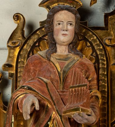 Zdjęcie nr 5: Rzeźba drążona, umieszczona w ołtarzu św. Mikołaja, na tle niszy lewego naddatku ołtarza. Święta Cecylia przedstawiona frontalnie w pozycji stojącej, w lekkim kontrapoście z lewą nogą lekko ugiętą i wysuniętą do przodu. Ukazana jako młoda, długowłosa kobieta o owalnej twarzy z pełnymi policzkami i niewielkim, wyraźnie zaznaczonym podbródkiem. Oczy o migdałowatym kształcie wpatrzone przed siebie, niewielkie usta zaznaczone czerwienią; mocno zaakcentowany, prosty nos. Głowę okalają długie, lekko falowane włosy opadające symetrycznie na ramiona postaci. Święta w lewej ręce trzyma złocony portatyw; prawa ręka zgięta w łokci i wysunięta nieco do przodu. Postać ubrana jest w długą, złotą suknię oraz szatę wierzchnią z długimi rękawami oraz trójkątnie wyciętym dekoltem wykończonym kołnierzem. Przez prawe przedramię postaci przewieszony fałd sukni. Spod sukni widoczne brązowe pantofle o miękko zakończonych noskach. Partie karnacji oraz włosów polichromowane, szaty i portatyw złocone.