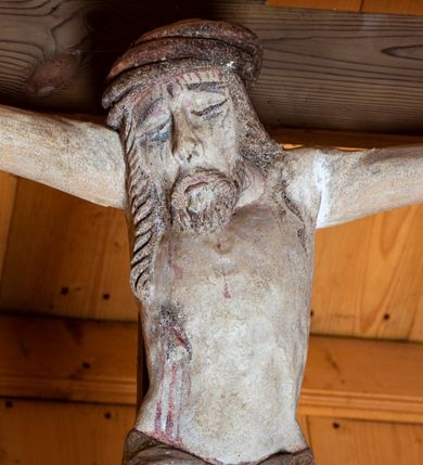 Zdjęcie nr 5: Chrystus ukazany w typie Cristo morte. Ciało przybite do prostego, drewnianego krzyża trzema gwoździami. Chrystus z szeroko rozpostartymi ramionami tworzącymi niemal linię prostą. Głowa przechylona nieznacznie w kierunku prawego barku. Ukazana w cierniowej koronie, spod której sączą się strużki krwi – nieproporcjonalnie duża w stosunku do wychudzonego ciała postaci. Twarz prostokątna z dużymi, zamkniętymi oczami, wydatnym nosem oraz ciemnym zarostem. Włosy długie, spływające na ramiona i plecy postaci w dwóch spiralnie skręconych puklach. Ciało Chrystusa szczupłe o wysmukłych proporcjach, bez wyraźnie zaznaczonej muskulatury. Dłonie, przebite gwoździami zamykają się lekko na wydatnych główkach gwoździ. Po przedramionach spływają cienkie stróżki krwi. Stopy duże, przebite jednym gwoździem. Na prawym boku postaci widoczna krwawiąca rana. Perizonium krótkie, srebrzone, o ostro łamiącej się draperii. Nad głową Chrystusa titulus ze złoconym napisem „INRI” wykonanym gotycką minuskułą.