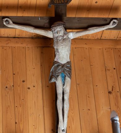 Zdjęcie nr 1: Chrystus ukazany w typie Cristo morte. Ciało przybite do prostego, drewnianego krzyża trzema gwoździami. Chrystus z szeroko rozpostartymi ramionami tworzącymi niemal linię prostą. Głowa przechylona nieznacznie w kierunku prawego barku. Ukazana w cierniowej koronie, spod której sączą się strużki krwi – nieproporcjonalnie duża w stosunku do wychudzonego ciała postaci. Twarz prostokątna z dużymi, zamkniętymi oczami, wydatnym nosem oraz ciemnym zarostem. Włosy długie, spływające na ramiona i plecy postaci w dwóch spiralnie skręconych puklach. Ciało Chrystusa szczupłe o wysmukłych proporcjach, bez wyraźnie zaznaczonej muskulatury. Dłonie, przebite gwoździami zamykają się lekko na wydatnych główkach gwoździ. Po przedramionach spływają cienkie stróżki krwi. Stopy duże, przebite jednym gwoździem. Na prawym boku postaci widoczna krwawiąca rana. Perizonium krótkie, srebrzone, o ostro łamiącej się draperii. Nad głową Chrystusa titulus ze złoconym napisem „INRI” wykonanym gotycką minuskułą.