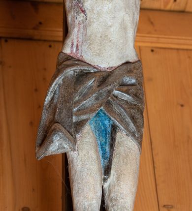 Zdjęcie nr 6: Chrystus ukazany w typie Cristo morte. Ciało przybite do prostego, drewnianego krzyża trzema gwoździami. Chrystus z szeroko rozpostartymi ramionami tworzącymi niemal linię prostą. Głowa przechylona nieznacznie w kierunku prawego barku. Ukazana w cierniowej koronie, spod której sączą się strużki krwi – nieproporcjonalnie duża w stosunku do wychudzonego ciała postaci. Twarz prostokątna z dużymi, zamkniętymi oczami, wydatnym nosem oraz ciemnym zarostem. Włosy długie, spływające na ramiona i plecy postaci w dwóch spiralnie skręconych puklach. Ciało Chrystusa szczupłe o wysmukłych proporcjach, bez wyraźnie zaznaczonej muskulatury. Dłonie, przebite gwoździami zamykają się lekko na wydatnych główkach gwoździ. Po przedramionach spływają cienkie stróżki krwi. Stopy duże, przebite jednym gwoździem. Na prawym boku postaci widoczna krwawiąca rana. Perizonium krótkie, srebrzone, o ostro łamiącej się draperii. Nad głową Chrystusa titulus ze złoconym napisem „INRI” wykonanym gotycką minuskułą.