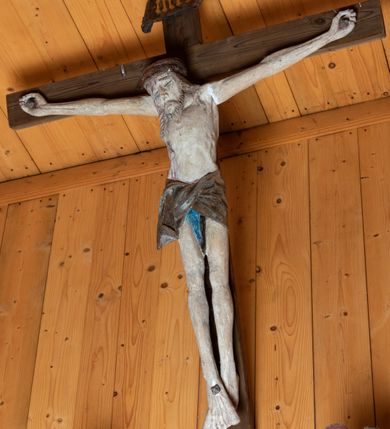 Zdjęcie nr 8: Chrystus ukazany w typie Cristo morte. Ciało przybite do prostego, drewnianego krzyża trzema gwoździami. Chrystus z szeroko rozpostartymi ramionami tworzącymi niemal linię prostą. Głowa przechylona nieznacznie w kierunku prawego barku. Ukazana w cierniowej koronie, spod której sączą się strużki krwi – nieproporcjonalnie duża w stosunku do wychudzonego ciała postaci. Twarz prostokątna z dużymi, zamkniętymi oczami, wydatnym nosem oraz ciemnym zarostem. Włosy długie, spływające na ramiona i plecy postaci w dwóch spiralnie skręconych puklach. Ciało Chrystusa szczupłe o wysmukłych proporcjach, bez wyraźnie zaznaczonej muskulatury. Dłonie, przebite gwoździami zamykają się lekko na wydatnych główkach gwoździ. Po przedramionach spływają cienkie stróżki krwi. Stopy duże, przebite jednym gwoździem. Na prawym boku postaci widoczna krwawiąca rana. Perizonium krótkie, srebrzone, o ostro łamiącej się draperii. Nad głową Chrystusa titulus ze złoconym napisem „INRI” wykonanym gotycką minuskułą.