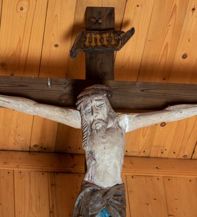 Zdjęcie nr 4: Chrystus ukazany w typie Cristo morte. Ciało przybite do prostego, drewnianego krzyża trzema gwoździami. Chrystus z szeroko rozpostartymi ramionami tworzącymi niemal linię prostą. Głowa przechylona nieznacznie w kierunku prawego barku. Ukazana w cierniowej koronie, spod której sączą się strużki krwi – nieproporcjonalnie duża w stosunku do wychudzonego ciała postaci. Twarz prostokątna z dużymi, zamkniętymi oczami, wydatnym nosem oraz ciemnym zarostem. Włosy długie, spływające na ramiona i plecy postaci w dwóch spiralnie skręconych puklach. Ciało Chrystusa szczupłe o wysmukłych proporcjach, bez wyraźnie zaznaczonej muskulatury. Dłonie, przebite gwoździami zamykają się lekko na wydatnych główkach gwoździ. Po przedramionach spływają cienkie stróżki krwi. Stopy duże, przebite jednym gwoździem. Na prawym boku postaci widoczna krwawiąca rana. Perizonium krótkie, srebrzone, o ostro łamiącej się draperii. Nad głową Chrystusa titulus ze złoconym napisem „INRI” wykonanym gotycką minuskułą.