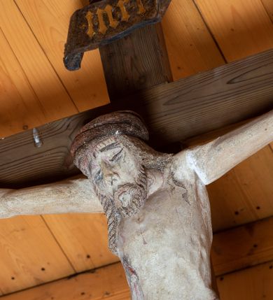 Zdjęcie nr 10: Chrystus ukazany w typie Cristo morte. Ciało przybite do prostego, drewnianego krzyża trzema gwoździami. Chrystus z szeroko rozpostartymi ramionami tworzącymi niemal linię prostą. Głowa przechylona nieznacznie w kierunku prawego barku. Ukazana w cierniowej koronie, spod której sączą się strużki krwi – nieproporcjonalnie duża w stosunku do wychudzonego ciała postaci. Twarz prostokątna z dużymi, zamkniętymi oczami, wydatnym nosem oraz ciemnym zarostem. Włosy długie, spływające na ramiona i plecy postaci w dwóch spiralnie skręconych puklach. Ciało Chrystusa szczupłe o wysmukłych proporcjach, bez wyraźnie zaznaczonej muskulatury. Dłonie, przebite gwoździami zamykają się lekko na wydatnych główkach gwoździ. Po przedramionach spływają cienkie stróżki krwi. Stopy duże, przebite jednym gwoździem. Na prawym boku postaci widoczna krwawiąca rana. Perizonium krótkie, srebrzone, o ostro łamiącej się draperii. Nad głową Chrystusa titulus ze złoconym napisem „INRI” wykonanym gotycką minuskułą.
