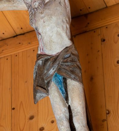 Zdjęcie nr 3: Chrystus ukazany w typie Cristo morte. Ciało przybite do prostego, drewnianego krzyża trzema gwoździami. Chrystus z szeroko rozpostartymi ramionami tworzącymi niemal linię prostą. Głowa przechylona nieznacznie w kierunku prawego barku. Ukazana w cierniowej koronie, spod której sączą się strużki krwi – nieproporcjonalnie duża w stosunku do wychudzonego ciała postaci. Twarz prostokątna z dużymi, zamkniętymi oczami, wydatnym nosem oraz ciemnym zarostem. Włosy długie, spływające na ramiona i plecy postaci w dwóch spiralnie skręconych puklach. Ciało Chrystusa szczupłe o wysmukłych proporcjach, bez wyraźnie zaznaczonej muskulatury. Dłonie, przebite gwoździami zamykają się lekko na wydatnych główkach gwoździ. Po przedramionach spływają cienkie stróżki krwi. Stopy duże, przebite jednym gwoździem. Na prawym boku postaci widoczna krwawiąca rana. Perizonium krótkie, srebrzone, o ostro łamiącej się draperii. Nad głową Chrystusa titulus ze złoconym napisem „INRI” wykonanym gotycką minuskułą.