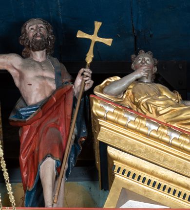 Zdjęcie nr 1: Pełnoplastyczna, polichromowana figura Chrystusa Zmartwychwstałego ustawiona na niewielkim postumencie. Chrystus przedstawiony w pozycji stojącej, frontalnie, w lekkim kontrapoście, z prawą nogą lekko ugiętą i wysuniętą nieco do przodu. Prawa ręka uniesiona do góry w geście błogosławieństwa. Lewa wsparta na złoconym krzyżu umieszczonym na długim drzewcu. Głowa nieznacznie przechylona w lewo; uniesiona ku górze. Chrystus ukazany jako dojrzały, brodaty mężczyzna o nagim torsie z widoczną raną w prawym boku. Wyraźnie zaznaczona muskulatura. Twarz szczupła, okolona półdługimi, falowanymi włosami w kolorze brązowym i takim samym zarostem. Przez lewe ramię postaci przewieszony czerwony płaszcz z ciemnogranatową podszewką oraz złotą lamówką. Draperia szat obficie fałduje się, tworząc głębokie, ostro łamiące się fałdy.
