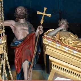 Zdjęcie nr 1: Pełnoplastyczna, polichromowana figura Chrystusa Zmartwychwstałego ustawiona na niewielkim postumencie. Chrystus przedstawiony w pozycji stojącej, frontalnie, w lekkim kontrapoście, z prawą nogą lekko ugiętą i wysuniętą nieco do przodu. Prawa ręka uniesiona do góry w geście błogosławieństwa. Lewa wsparta na złoconym krzyżu umieszczonym na długim drzewcu. Głowa nieznacznie przechylona w lewo; uniesiona ku górze. Chrystus ukazany jako dojrzały, brodaty mężczyzna o nagim torsie z widoczną raną w prawym boku. Wyraźnie zaznaczona muskulatura. Twarz szczupła, okolona półdługimi, falowanymi włosami w kolorze brązowym i takim samym zarostem. Przez lewe ramię postaci przewieszony czerwony płaszcz z ciemnogranatową podszewką oraz złotą lamówką. Draperia szat obficie fałduje się, tworząc głębokie, ostro łamiące się fałdy.
