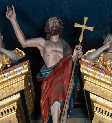 Zdjęcie nr 2: Pełnoplastyczna, polichromowana figura Chrystusa Zmartwychwstałego ustawiona na niewielkim postumencie. Chrystus przedstawiony w pozycji stojącej, frontalnie, w lekkim kontrapoście, z prawą nogą lekko ugiętą i wysuniętą nieco do przodu. Prawa ręka uniesiona do góry w geście błogosławieństwa. Lewa wsparta na złoconym krzyżu umieszczonym na długim drzewcu. Głowa nieznacznie przechylona w lewo; uniesiona ku górze. Chrystus ukazany jako dojrzały, brodaty mężczyzna o nagim torsie z widoczną raną w prawym boku. Wyraźnie zaznaczona muskulatura. Twarz szczupła, okolona półdługimi, falowanymi włosami w kolorze brązowym i takim samym zarostem. Przez lewe ramię postaci przewieszony czerwony płaszcz z ciemnogranatową podszewką oraz złotą lamówką. Draperia szat obficie fałduje się, tworząc głębokie, ostro łamiące się fałdy.
