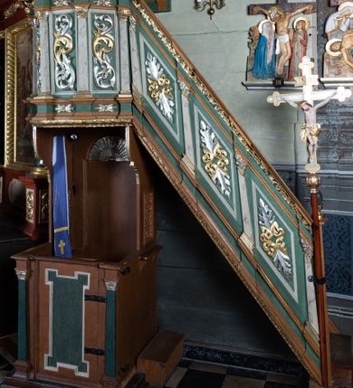 Zdjęcie nr 1: Ambona przyścienna o formach architektonicznych; dostępna jednobiegowymi schodkami od strony prezbiterium. Kosz na rzucie wieloboku, ustawiony na znajdującym się poniżej konfesjonale, ujęty pilastrami o kompozytowych, złoconych kapitelach. Pilastry stojące na wysuniętych przed lico ambony profilowanych postumentach dźwigają wydatny, profilowany i wyłamujący się na osi podpór gzyms. Prostokątne pola kosza są wypełnione złoconym i srebrzonym „suchym” akantem. Fryz i architraw dekorowane złoconym ornamentem kwiatowym. Balustrada schodków pełna o podziałach identycznych jak korpus ambony. Zaplecek ujęty parą pilastrów o kompozytowych, złoconych kapitelach dźwigających pełne, wyłamane na osi podpór belkowanie zwieńczone profilowanym gzymsem. W polu obraz przedstawiający św. Franciszka z Asyżu na tle pejzażu w kształcie stojącego prostokąta zamkniętego łukiem nadwieszonym. Święty został ukazany całopostaciowo, frontalnie w brązowym habicie przepasanym sznurem. W lewej dłoni trzyma zamkniętą księgę oraz lilię, prawą wykonuje gest oratorski. U jego lewego boku przedstawiony jeleń. Całość zaplecka ujęta ażurowymi uszami utworzonymi ze złoconego i srebrzonego „suchego” akantu. Baldachim na rzucie połowy ośmioramiennej gwiazdy, zwieńczony złoconą szyszką ustawioną na ażurowej konsoli utworzonej z akantowych wolut; w podniebiu płaskorzeźbiona gołębica Ducha Świętego na tle glorii promienistej. Struktura malowana na kolor jasnozielony z jasnobrązowymi elementami. Partie ornamentów złocone i srebrzone; trzony pilastrów marmoryzowane w odcieniach pastelowej zieleni. Konfesjonał na rzucie prostokąta. Przedpiersie ujęte parą pilastrów o gładkich, ciemnozielonych trzonach i złoconych, kompozytowych kapitelach. W polu prostokątna, „uszata” płycina malowana na ciemnozielono. Zaplecek gładki z prostokątną płyciną zamkniętą łukiem nadwieszonym mieszczącym złoconą muszlę palmetową. Boki o linii wklęsło-wypukłej zamknięte od góry akantowymi wolutami. Struktura malowana na kolor jasnobrązowy.

