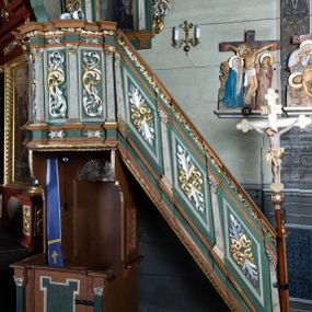 Zdjęcie nr 1: Ambona przyścienna o formach architektonicznych; dostępna jednobiegowymi schodkami od strony prezbiterium. Kosz na rzucie wieloboku, ustawiony na znajdującym się poniżej konfesjonale, ujęty pilastrami o kompozytowych, złoconych kapitelach. Pilastry stojące na wysuniętych przed lico ambony profilowanych postumentach dźwigają wydatny, profilowany i wyłamujący się na osi podpór gzyms. Prostokątne pola kosza są wypełnione złoconym i srebrzonym „suchym” akantem. Fryz i architraw dekorowane złoconym ornamentem kwiatowym. Balustrada schodków pełna o podziałach identycznych jak korpus ambony. Zaplecek ujęty parą pilastrów o kompozytowych, złoconych kapitelach dźwigających pełne, wyłamane na osi podpór belkowanie zwieńczone profilowanym gzymsem. W polu obraz przedstawiający św. Franciszka z Asyżu na tle pejzażu w kształcie stojącego prostokąta zamkniętego łukiem nadwieszonym. Święty został ukazany całopostaciowo, frontalnie w brązowym habicie przepasanym sznurem. W lewej dłoni trzyma zamkniętą księgę oraz lilię, prawą wykonuje gest oratorski. U jego lewego boku przedstawiony jeleń. Całość zaplecka ujęta ażurowymi uszami utworzonymi ze złoconego i srebrzonego „suchego” akantu. Baldachim na rzucie połowy ośmioramiennej gwiazdy, zwieńczony złoconą szyszką ustawioną na ażurowej konsoli utworzonej z akantowych wolut; w podniebiu płaskorzeźbiona gołębica Ducha Świętego na tle glorii promienistej. Struktura malowana na kolor jasnozielony z jasnobrązowymi elementami. Partie ornamentów złocone i srebrzone; trzony pilastrów marmoryzowane w odcieniach pastelowej zieleni. Konfesjonał na rzucie prostokąta. Przedpiersie ujęte parą pilastrów o gładkich, ciemnozielonych trzonach i złoconych, kompozytowych kapitelach. W polu prostokątna, „uszata” płycina malowana na ciemnozielono. Zaplecek gładki z prostokątną płyciną zamkniętą łukiem nadwieszonym mieszczącym złoconą muszlę palmetową. Boki o linii wklęsło-wypukłej zamknięte od góry akantowymi wolutami. Struktura malowana na kolor jasnobrązowy.
