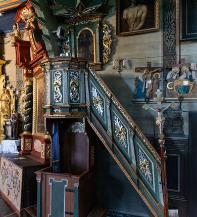 Zdjęcie nr 3: Ambona przyścienna o formach architektonicznych; dostępna jednobiegowymi schodkami od strony prezbiterium. Kosz na rzucie wieloboku, ustawiony na znajdującym się poniżej konfesjonale, ujęty pilastrami o kompozytowych, złoconych kapitelach. Pilastry stojące na wysuniętych przed lico ambony profilowanych postumentach dźwigają wydatny, profilowany i wyłamujący się na osi podpór gzyms. Prostokątne pola kosza są wypełnione złoconym i srebrzonym „suchym” akantem. Fryz i architraw dekorowane złoconym ornamentem kwiatowym. Balustrada schodków pełna o podziałach identycznych jak korpus ambony. Zaplecek ujęty parą pilastrów o kompozytowych, złoconych kapitelach dźwigających pełne, wyłamane na osi podpór belkowanie zwieńczone profilowanym gzymsem. W polu obraz przedstawiający św. Franciszka z Asyżu na tle pejzażu w kształcie stojącego prostokąta zamkniętego łukiem nadwieszonym. Święty został ukazany całopostaciowo, frontalnie w brązowym habicie przepasanym sznurem. W lewej dłoni trzyma zamkniętą księgę oraz lilię, prawą wykonuje gest oratorski. U jego lewego boku przedstawiony jeleń. Całość zaplecka ujęta ażurowymi uszami utworzonymi ze złoconego i srebrzonego „suchego” akantu. Baldachim na rzucie połowy ośmioramiennej gwiazdy, zwieńczony złoconą szyszką ustawioną na ażurowej konsoli utworzonej z akantowych wolut; w podniebiu płaskorzeźbiona gołębica Ducha Świętego na tle glorii promienistej. Struktura malowana na kolor jasnozielony z jasnobrązowymi elementami. Partie ornamentów złocone i srebrzone; trzony pilastrów marmoryzowane w odcieniach pastelowej zieleni. Konfesjonał na rzucie prostokąta. Przedpiersie ujęte parą pilastrów o gładkich, ciemnozielonych trzonach i złoconych, kompozytowych kapitelach. W polu prostokątna, „uszata” płycina malowana na ciemnozielono. Zaplecek gładki z prostokątną płyciną zamkniętą łukiem nadwieszonym mieszczącym złoconą muszlę palmetową. Boki o linii wklęsło-wypukłej zamknięte od góry akantowymi wolutami. Struktura malowana na kolor jasnobrązowy.
