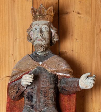 Zdjęcie nr 2: Rzeźba pełnoplastyczna, ścięta z tyłu. Król ukazany w postawie stojącej, frontalnie, z nogami w lekkim rozkroku i ugiętymi w kolanach. Ręce zgięte w łokciach pod kątem prostym z ramionami przylegającymi do tułowia. Przedramiona skierowane w lewo. Głowa wydłużona, jasnobrązowe włosy dłuższe, lekko falowane, odsłaniające uszy, zarost krótki. Wzrok skierowany przed siebie, oczy duże migdałowate, brwi wyraźne, łukowe. Nos długi, wydatny, usta niewielkie. Na głowie postaci złocona korona w typie otwartym. Król ubrany jest w zbroję, na którą składa się krótki, łuskowy pancerz, zarękawia, nabiodrki, nakolanki, nagolennice i trzewiki. Na pancerz narzucony jest długi, złoty płaszcz, podszyty na czerwono, spięty pod szyją okrągłą zapinką. Figura w pełni polichromowana, korona i płaszcz złocone.