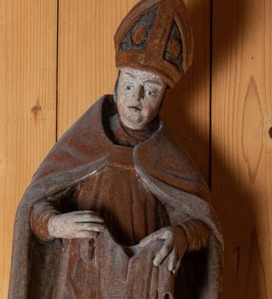 Zdjęcie nr 2: Rzeźba pełnoplastyczna, ścięta z tyłu, ustawiona na niewielkiej podstawie o nieregularnym kształcie. Biskup ukazany w całej postaci, frontalnie w postawie stojącej. U jego stóp po prawej stronie kompozycji przedstawiona w pomniejszonej skali postać klęczącego żebraka. Głowa św. Marcina lekko pochylona do dołu. Święty w ugiętych łokciach rękach trzyma przed sobą szatę, którą zamierza nałożyć klęczącemu żebrakowi. Twarz biskupa podłużna o wyraźnie zaznaczonym, lekko zaokrąglonym podbródku, krótkim nosie oraz lekko rozchylonych ustach; pozbawiona zarostu. Biskup ubrany jest w długą albę, złoconą dalmatykę sięgającą za kolana oraz złoty, podbity srebrną tkaniną płaszcz sięgający ziemi. Na głowie świętego przedstawiona infuła biskupia. Na dłoniach rękawiczki. Spod alby widoczna prawa stopa w obuwiu o zaokrąglonych noskach. Żebrak przedstawiony jako brodaty mężczyzna o okrągłej, pozbawionej włosów głowie. Swój wzrok kieruje ku górze, w kierunku św. Marcina. Prawą, uniesioną do góry ręką chwyta brzeg szaty nakładanej mu przez świętego. Lewa ręka, wsparta na kuli, opuszczona wzdłuż tułowia. Przez prawe ramię  postaci przewieszona srebrna szata, luźno okrywająca ciało. Tors odsłonięty.

