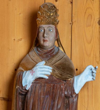 Zdjęcie nr 2: Rzeźba pełnoplastyczna, ścięta z tyłu, ustawiona na niewielkim postumencie. Święty ukazany w całej postaci, stojący frontalnie, w delikatnym kontrapoście, z lewą nogą lekko ugiętą w kolanie. Ręce postaci zgięte w łokciach. Prawa dłoń świętego spoczywa na piersi, lewa odsunięta jest w bok od ciała. Twarz papieża okrągła, pozbawiona zarostu, z pełnymi policzkami oraz podwójnym podbródkiem. Oczy duże migdałowate, wzrok skierowany przed siebie. Brwi wyraźne, łukowate, nos długi, prosty, usta niewielkie. Na głowie świętego umieszczona złota tiara. Papież ubrany jest w długą, czerwoną szatę oraz złotą, spiętą pod szyją kapę, której brzeg dekorowany jest wzorem z rombów. Na rękach postaci białe, sięgające łokci rękawice dekorowane czerwonymi krzyżami. Spod szaty widoczne czubki butów.