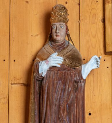 Zdjęcie nr 1: Rzeźba pełnoplastyczna, ścięta z tyłu, ustawiona na niewielkim postumencie. Święty ukazany w całej postaci, stojący frontalnie, w delikatnym kontrapoście, z lewą nogą lekko ugiętą w kolanie. Ręce postaci zgięte w łokciach. Prawa dłoń świętego spoczywa na piersi, lewa odsunięta jest w bok od ciała. Twarz papieża okrągła, pozbawiona zarostu, z pełnymi policzkami oraz podwójnym podbródkiem. Oczy duże migdałowate, wzrok skierowany przed siebie. Brwi wyraźne, łukowate, nos długi, prosty, usta niewielkie. Na głowie świętego umieszczona złota tiara. Papież ubrany jest w długą, czerwoną szatę oraz złotą, spiętą pod szyją kapę, której brzeg dekorowany jest wzorem z rombów. Na rękach postaci białe, sięgające łokci rękawice dekorowane czerwonymi krzyżami. Spod szaty widoczne czubki butów.