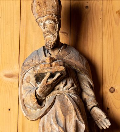 Zdjęcie nr 2: Figura drążona, ustawiona na niewielkiej podstawie. Święty został ukazany w całej postaci, w postawie stojącej, w wyraźnym kontrapoście, z prawą nogą wysuniętą do przodu. Głowa postaci zwrócona jest w trzech czwartych w prawo, wzrok skierowany przed siebie. Twarz świętego szczupła, pociągła z krótkim nosem, niewielkimi ustami oraz kędzierzawą brodą. W prawej zgiętej w łokciu ręce święty podtrzymuje otwartą księgę, na której znajdują się trzy kule. Lewa ręka postaci opuszczona wzdłuż tułowia. Święty ubrany jest w długą, obficie fałdowaną suknię oraz spięty pod szyją płaszcz. Na jego głowie znajduje się infuła biskupia. 