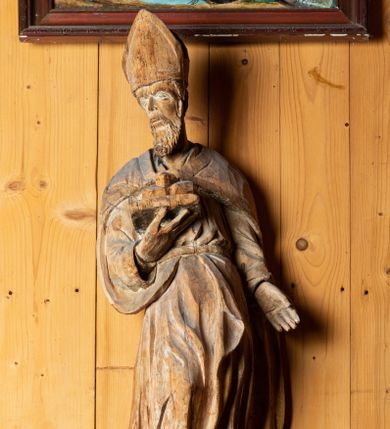 Zdjęcie nr 1: Figura drążona, ustawiona na niewielkiej podstawie. Święty został ukazany w całej postaci, w postawie stojącej, w wyraźnym kontrapoście, z prawą nogą wysuniętą do przodu. Głowa postaci zwrócona jest w trzech czwartych w prawo, wzrok skierowany przed siebie. Twarz świętego szczupła, pociągła z krótkim nosem, niewielkimi ustami oraz kędzierzawą brodą. W prawej zgiętej w łokciu ręce święty podtrzymuje otwartą księgę, na której znajdują się trzy kule. Lewa ręka postaci opuszczona wzdłuż tułowia. Święty ubrany jest w długą, obficie fałdowaną suknię oraz spięty pod szyją płaszcz. Na jego głowie znajduje się infuła biskupia. 