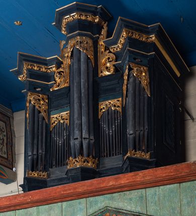 Zdjęcie nr 2: Prospekt organowy o formach architektonicznych, pięcioosiowy, jednokondygnacyjny ze zwieńczeniem. Osie nieparzyste wypukłe; osie skrajne na rzucie trójkąta; oś środkowa wyższa na rzucie połowy sześcioboku ujęta w górnej części akantowymi wolutami. W osiach parzystych niższe płyciny mieszczące mniejsze piszczałki. W górnej partii części piszczałkowej płaskorzeźbiona dekoracja akantowa. Zwieńczenie utworzone z przełamującego się, bogato profilowanego belkowania z wydatnym gzymsem. Struktura malowana w kolorze ciemnego granatu, ornamenty w jaśniejszym zielono-niebieskim odcieniu.