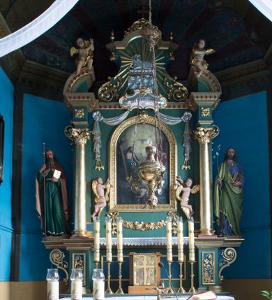 Zdjęcie nr 1: Ołtarz architektoniczny, jednokondygnacyjny, jednoosiowy ze zwieńczeniem. Ołtarz prostopadłościenny, z drewnianą mensą. Antepedium ujęte szerokim obramieniem, pośrodku w polu w kształcie koła złocony monogram imienia Jezus. Retabulum osadzone na zryzalitowanym po bokach cokole, ujętym parą wolutowych impostów. Część środkowa flankowana parą kolumn wspierających niepełne, wyłamujące się ponad nimi, przerwane w partii fryzu belkowanie. Zwieńczenie ujęte parą spływów wolutowych, zamknięte fragmentem gzymsu w postaci półkolistego łuku nadwieszonego, po bokach przerwany, półkolisty przyczółek. Pola cokołu zdobione motywami wici roślinnej. W dolnej części pola środkowego nastawy kwietna girlanda, wyżej, po bokach para aniołków, w górnej partii pośrodku gzymsu kartusz z napisem „OTO BARANEK BOŻY” z podwieszeniem w formie podwiązanej w narożach polach kotary. W partiach fryzu rozety. W zwieńczeniu motyw płaskorzeźbionej Baranka Paschalnego ujętego promienistą glorią. Dolną część spływów wolutowych zdobią liście akantu. Szczyt w formie krzyża osadzonego na ażurowej podstawie w postaci wici akantowej, ujęty parą płomienistych wazonów. Na przerwanym przyczółku para figur siedzących aniołków. Pole części środkowej retabulum w kształcie zbliżonym do kwadratu, zamkniętym półkoliście, ujętym rzeźbioną, złoconą ramą. W środku obraz „Trójcy Świętej”. Po bokach retabulum para rzeźb, po lewej św. Jakub, po prawej św. Józef. Struktura polichromowana w kolorze zielonym i niebieskim, detal architektoniczny złocony i srebrzony.   