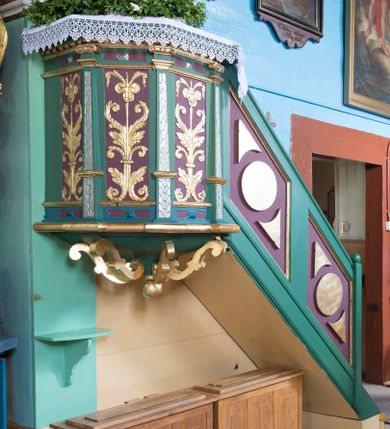 Zdjęcie nr 1: Ambona przyścienna, wisząca, z wejściem schodami od strony prezbiterium. Kosz jest na planie pięcioboku z ośmioboku ze ściankami wydzielonymi wąskimi pilastrami na wysokich cokołach, o trzonach zdobionych motywem liści laurowych. Pola ścianek wypełniają płyciny w formie stojącego prostokąta pokryte ornamentem kandelabrowym. Kosz dołem obiega gzyms w kształcie półwałka. Ujęty ornamentem sznurowym parapet jest w formie gzymsu. Spód kosza w postaci wklęsło-wypukłego, zdobionego czterema kabłąkami w formie S-kształtnych wolut, ostrosłupa  z podwieszeniem w formie kuli. Schody są jednobiegowe z balustradą w kształcie ścianki wypełnioną parą romboidalnych płycin z motywem koła i pary trójkątów o wklęsłej podstawie. Struktura polichromowana w kolorach zielonym, niebieskim i fioletowym, detal architektoniczny i ornamentyka złocone i srebrzone.  