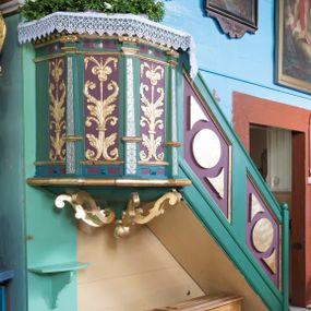 Zdjęcie nr 1: Ambona przyścienna, wisząca, z wejściem schodami od strony prezbiterium. Kosz jest na planie pięcioboku z ośmioboku ze ściankami wydzielonymi wąskimi pilastrami na wysokich cokołach, o trzonach zdobionych motywem liści laurowych. Pola ścianek wypełniają płyciny w formie stojącego prostokąta pokryte ornamentem kandelabrowym. Kosz dołem obiega gzyms w kształcie półwałka. Ujęty ornamentem sznurowym parapet jest w formie gzymsu. Spód kosza w postaci wklęsło-wypukłego, zdobionego czterema kabłąkami w formie S-kształtnych wolut, ostrosłupa  z podwieszeniem w formie kuli. Schody są jednobiegowe z balustradą w kształcie ścianki wypełnioną parą romboidalnych płycin z motywem koła i pary trójkątów o wklęsłej podstawie. Struktura polichromowana w kolorach zielonym, niebieskim i fioletowym, detal architektoniczny i ornamentyka złocone i srebrzone.  