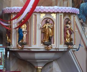 Zdjęcie nr 1: Ambona przyścienna, wisząca, z dostępem w postaci jednobiegowych schodów z pełną balustradą, dostawionych od północy. Kosz na planie ośmioboku, ustawiony na niskim, profilowanym cokole, z podwieszeniem w formie zbliżonej do spłaszczonego stożka, o wklęsło-wypukłych ściankach, zakończonego szyszką. Ścianki ambony wydzielone zwielokrotnionymi pilastrami toskańskimi, z płycinami w partii trzonów, podtrzymującymi przełamujący się nad nimi gzyms tworzący parapet. W polach płyciny w formie stojącego prostokąta zamkniętego łukiem półkolistym z figurami czterech ewangelistów ustawionych na wspornikach w postaci uskrzydlonych główek anielskich. Baldachim na planie ośmioboku, w formie zbliżonej do spłaszczonego stożka, o wklęsło-wypukłych ściankach, o krawędzi obwiedzionej lambrekinem, zakończonego postumentem z figurą Chrystusa w typie Salvator Mundi. Na podniebieniu gołębica Ducha Świętego w glorii promienistej. Struktura polichromowana w odcieniach beżu, z bladoróżowymi detalami i złoconymi profilami. Figury ewangelistów i Chrystusa ścięte z tyłu, ukazane frontalnie, w kontrapostach. Twarze mają pociągłe, o migdałowatych oczach, garbatych nosach i pełnych ustach, okolone są gęstymi, krótkimi brodami i falowanymi włosami opadającymi na ramiona. Postacie ubrane są w złocone tuniki przepasane w talii oraz płaszcze. Ewangeliści trzymają w rękach pióra i otwarte księgi, przy nogach znajdują się ich atrybuty. Chrystus w lewej ręce trzyma jabłko królewskie, prawą błogosławi. 