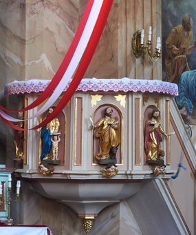Zdjęcie nr 1: Ambona przyścienna, wisząca, z dostępem w postaci jednobiegowych schodów z pełną balustradą, dostawionych od północy. Kosz na planie ośmioboku, ustawiony na niskim, profilowanym cokole, z podwieszeniem w formie zbliżonej do spłaszczonego stożka, o wklęsło-wypukłych ściankach, zakończonego szyszką. Ścianki ambony wydzielone zwielokrotnionymi pilastrami toskańskimi, z płycinami w partii trzonów, podtrzymującymi przełamujący się nad nimi gzyms tworzący parapet. W polach płyciny w formie stojącego prostokąta zamkniętego łukiem półkolistym z figurami czterech ewangelistów ustawionych na wspornikach w postaci uskrzydlonych główek anielskich. Baldachim na planie ośmioboku, w formie zbliżonej do spłaszczonego stożka, o wklęsło-wypukłych ściankach, o krawędzi obwiedzionej lambrekinem, zakończonego postumentem z figurą Chrystusa w typie Salvator Mundi. Na podniebieniu gołębica Ducha Świętego w glorii promienistej. Struktura polichromowana w odcieniach beżu, z bladoróżowymi detalami i złoconymi profilami. Figury ewangelistów i Chrystusa ścięte z tyłu, ukazane frontalnie, w kontrapostach. Twarze mają pociągłe, o migdałowatych oczach, garbatych nosach i pełnych ustach, okolone są gęstymi, krótkimi brodami i falowanymi włosami opadającymi na ramiona. Postacie ubrane są w złocone tuniki przepasane w talii oraz płaszcze. Ewangeliści trzymają w rękach pióra i otwarte księgi, przy nogach znajdują się ich atrybuty. Chrystus w lewej ręce trzyma jabłko królewskie, prawą błogosławi. 