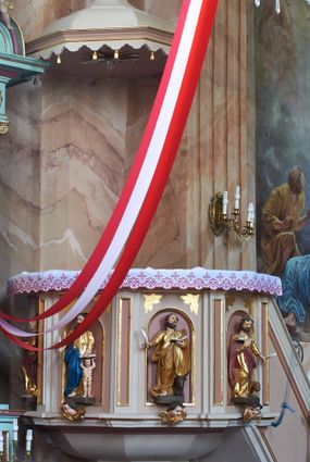 Zdjęcie nr 1: Ambona przyścienna, wisząca, z dostępem w postaci jednobiegowych schodów z pełną balustradą, dostawionych od północy. Kosz na planie ośmioboku, ustawiony na niskim, profilowanym cokole, z podwieszeniem w formie zbliżonej do spłaszczonego stożka, o wklęsło-wypukłych ściankach, zakończonego szyszką. Ścianki ambony wydzielone zwielokrotnionymi pilastrami toskańskimi, z płycinami w partii trzonów, podtrzymującymi przełamujący się nad nimi gzyms tworzący parapet. W polach płyciny w formie stojącego prostokąta zamkniętego łukiem półkolistym z figurami czterech ewangelistów ustawionych na wspornikach w postaci uskrzydlonych główek anielskich. Baldachim na planie ośmioboku, w formie zbliżonej do spłaszczonego stożka, o wklęsło-wypukłych ściankach, o krawędzi obwiedzionej lambrekinem, zakończonego postumentem z figurą Chrystusa w typie Salvator Mundi. Na podniebieniu gołębica Ducha Świętego w glorii promienistej. Struktura polichromowana w odcieniach beżu, z bladoróżowymi detalami i złoconymi profilami. Figury ewangelistów i Chrystusa ścięte z tyłu, ukazane frontalnie, w kontrapostach. Twarze mają pociągłe, o migdałowatych oczach, garbatych nosach i pełnych ustach, okolone są gęstymi, krótkimi brodami i falowanymi włosami opadającymi na ramiona. Postacie ubrane są w złocone tuniki przepasane w talii oraz płaszcze. Ewangeliści trzymają w rękach pióra i otwarte księgi, przy nogach znajdują się ich atrybuty. Chrystus w lewej ręce trzyma jabłko królewskie, prawą błogosławi. 