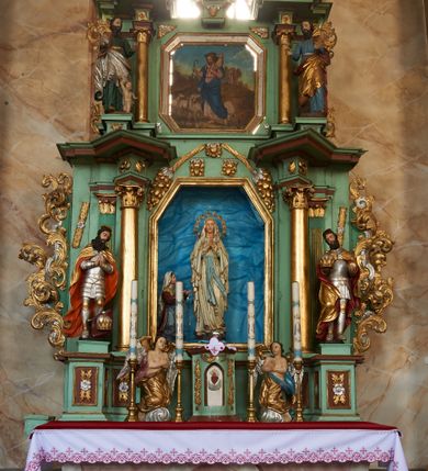 Zdjęcie nr 3: Nastawa ołtarzowa architektoniczna, jednoosiowa, dwukondygnacyjna na cokole z wyodrębnionymi dwoma prostopadłościanami. Ołtarz prostopadłościenny, zwężający się ku dołowi, mensa drewniana. Antepedium marmoryzowane z iluzjonistycznie malowanymi płycinami. Tabernakulum w formie drewnianej skrzynki, półkoliście wybrzuszone od frontu, zakończone profilowanym gzymsem. Drzwiczki o kształcie stojącego prostokąta zamknięte ostrołukiem z malowanym gorejącym sercem, flankowane płaskorzeźbioną winną latoroślą. Tabernakulum ujęte dwiema figurami klęczących na obłokach aniołów. Pierwsza kondygnacja flankowana dwiema parami pilastrów, zewnętrzna o kanelowanych trzonach. Przed wewnętrzną parą stoją dwie kolumny o kapitelach kompozytowych, przed zewnętrzną ustawione są figury św. Stefana i św. Władysława. Podpory dźwigają pełne belkowanie z silnie wysuniętym gzymsem, wyłamane pod kątem w partii kolumn, przerwane pośrodku. W polu centralnym nisza w formie stojącego prostokąta o ściętych górnych narożach ujęta profilowaną ramą, z grupą rzeźbiarską Matki Boskiej z Lourdes i Bernadetą Soubirous. W górnej partii nisza dekorowana zwisami owocowo-kwiatowymi. Pierwsza kondygnacja ujęta uszami utworzonymi z akantu i karbowanej wstęgi. Druga kondygnacja flankowana parą kolumn oraz parą figur św. Marka i św. Mateusza na cokołach. Podpory dźwigają pełne belkowanie z silnie wysuniętym gzymsem, wyłamane na ich osi, przerwane nad polem środkowym. Na belkowaniu dwie figurki aniołków, pośrodku na wsporniku figura Chrystusa w glorii, pośród obłoków. W polu obraz Chrystusa Dobrego Pasterza w formie leżącego prostokąta o ściętych narożach, w ramie. Druga kondygnacja ujęta uszami utworzonymi z akantu. Struktura polichromowana w kolorze jasnozielonym, kolumny, ornamentyka i profile złocone, detal srebrzony. 

