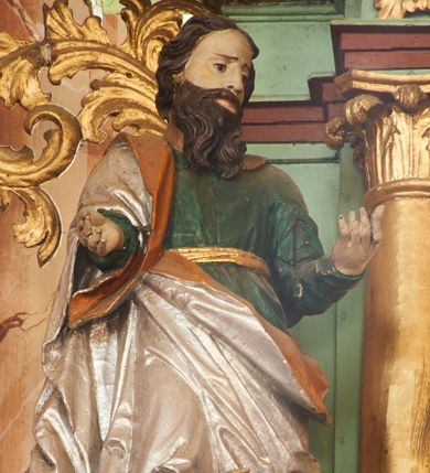 Zdjęcie nr 1: Święty ukazany frontalnie, z głową skierowaną w lewo, w kontrapoście, obie ręce ma ugięte, wyciągnięte do przodu. Twarz ma pociągłą, o małych oczach, ściągniętych brwiach, dużym nosie, szerokich ustach, okoloną długą brodą opadającą na pierś oraz krótkimi ciemnobrązowymi włosami. Ubrany jest w zieloną tunikę, przepasaną w talii złotą szarfą oraz srebrzony płaszcz z pomarańczową podszewką, zarzucony na prawe ramię i otaczający postać w partii bioder. Przy lewej nodze świętego postać klęczącego putta, unoszącego w prawej ręce dzbanuszek. Polichromia ciała figur naturalistyczna. 


