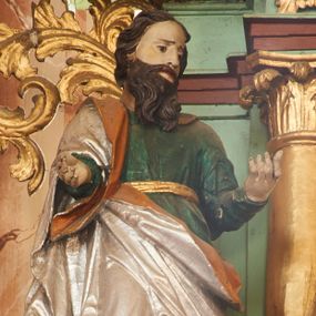 Zdjęcie nr 1: Święty ukazany frontalnie, z głową skierowaną w lewo, w kontrapoście, obie ręce ma ugięte, wyciągnięte do przodu. Twarz ma pociągłą, o małych oczach, ściągniętych brwiach, dużym nosie, szerokich ustach, okoloną długą brodą opadającą na pierś oraz krótkimi ciemnobrązowymi włosami. Ubrany jest w zieloną tunikę, przepasaną w talii złotą szarfą oraz srebrzony płaszcz z pomarańczową podszewką, zarzucony na prawe ramię i otaczający postać w partii bioder. Przy lewej nodze świętego postać klęczącego putta, unoszącego w prawej ręce dzbanuszek. Polichromia ciała figur naturalistyczna. 


