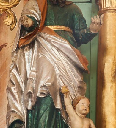 Zdjęcie nr 2: Święty ukazany frontalnie, z głową skierowaną w lewo, w kontrapoście, obie ręce ma ugięte, wyciągnięte do przodu. Twarz ma pociągłą, o małych oczach, ściągniętych brwiach, dużym nosie, szerokich ustach, okoloną długą brodą opadającą na pierś oraz krótkimi ciemnobrązowymi włosami. Ubrany jest w zieloną tunikę, przepasaną w talii złotą szarfą oraz srebrzony płaszcz z pomarańczową podszewką, zarzucony na prawe ramię i otaczający postać w partii bioder. Przy lewej nodze świętego postać klęczącego putta, unoszącego w prawej ręce dzbanuszek. Polichromia ciała figur naturalistyczna. 


