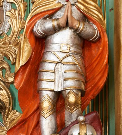 Zdjęcie nr 2: Święty ukazany frontalnie, z głową lekko przechyloną na lewe ramię, dłońmi złożonymi w geście modlitwy. Twarz ma pociągłą, o wąskich oczach, zadartym nosie i otwartych, małych ustach, okoloną długą brodą opadającą na pierś oraz krótkimi, ciemnymi włosami. Nad głową nimb. Ubrany jest w srebrzoną zbroję płytową o złoconych detalach, na ramiona ma narzucony złocony płaszcz o pomarańczowej podszewce, zapięty ozdobną klamrą na piersi. Na lewej stopie świętego leży czerwona poduszka z zamkniętą koroną. Polichromia ciała naturalistyczna.

