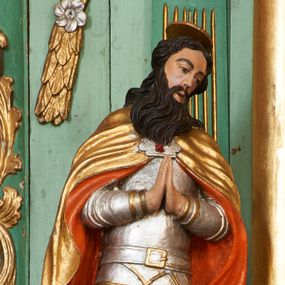 Zdjęcie nr 1: Święty ukazany frontalnie, z głową lekko przechyloną na lewe ramię, dłońmi złożonymi w geście modlitwy. Twarz ma pociągłą, o wąskich oczach, zadartym nosie i otwartych, małych ustach, okoloną długą brodą opadającą na pierś oraz krótkimi, ciemnymi włosami. Nad głową nimb. Ubrany jest w srebrzoną zbroję płytową o złoconych detalach, na ramiona ma narzucony złocony płaszcz o pomarańczowej podszewce, zapięty ozdobną klamrą na piersi. Na lewej stopie świętego leży czerwona poduszka z zamkniętą koroną. Polichromia ciała naturalistyczna.

